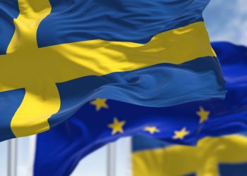 Detail of the national flag of Sweden waving in the wind with blurred european union flag in the background on a clear day. Democracy and politics. European country. Selective focus.